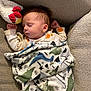 baby, sleeping, blanket, dinosaur_pattern, plush_toy, couch, cozy, child, pacifier, cute, infant, rest, soft_toy, fabric, comfort, relaxation, indoor, clothing, head, peaceful