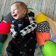 Quinn joined the competition — help win amazing prizes! baby, infant, smiling, happy, overalls, plaid_shirt, colorful_cushion, toy, stuffed_animal, soft_toy, floor_rug, child, play_area, indoor, cute, portrait, clothing, person, cozy, lying_down