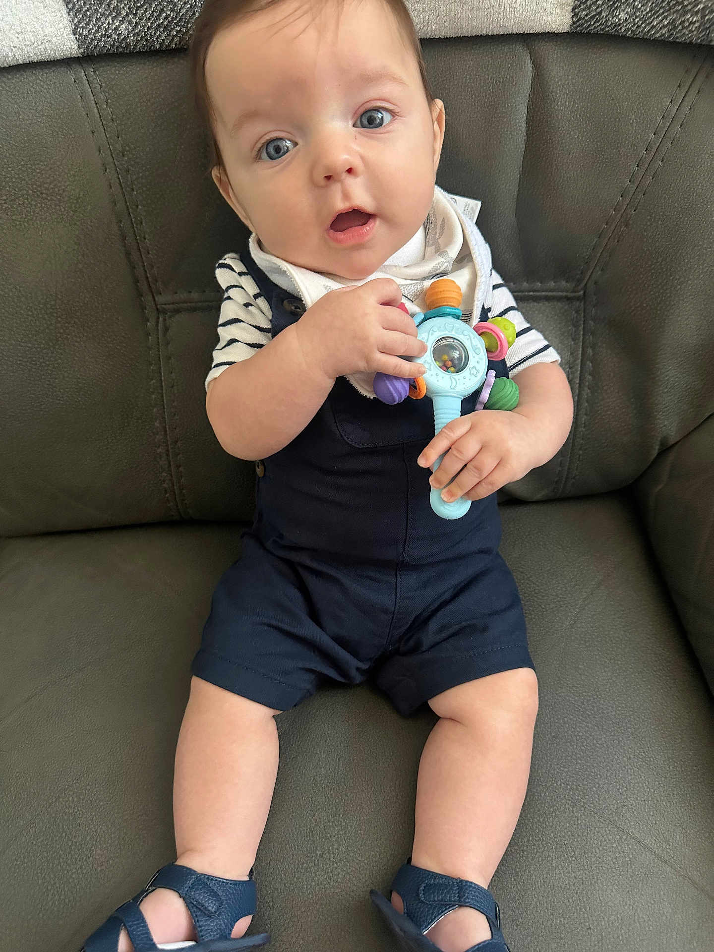Oscar joined the competition — help win amazing prizes! baby, child, infant, toy, blue_eyes, overalls, striped_shirt, sandals, leather_couch, curious, sitting, hands, legs, face, expression, indoor, comfortable, playful, closeup, person