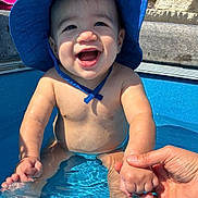 Lucian is registered to the contest to win money with this photo: baby, blue_hat, child, concrete, daytime, flower, foot, hand_holding, happy, headwear, outdoor, person, play, pool, skin, smiling, splashing, summer, sunny, water