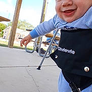 Lucian is registered to the contest to win money with this photo: baby, building, child, clothing, concrete, daylight, face, footwear, grass, hand, happy, harness, metal, outdoor, pajamas, person, picnic_table, sky, smiling, wood