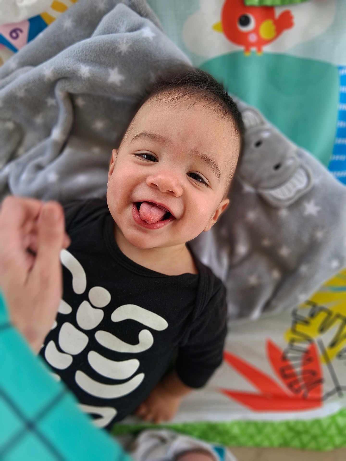 Lucian is registered to the contest to win money with this photo: baby, black_clothing, blanket, child, colorful, cute, face, happy, head, hippo, indoors, infant, person, play_mat, plush_toy, skeleton_print, smiling, toddler, tongue_out, young_child