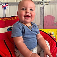 Maximo is registered to the contest to win money with this photo: baby, child, smile, blue_shirt, pants, sitting, cushion, playpen, indoor, happy, face, hand, foot, necklace, toy, furniture, colorful, person, young_child, closeup