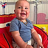 baby, child, smile, blue_shirt, pants, sitting, cushion, playpen, indoor, happy, face, hand, foot, necklace, toy, furniture, colorful, person, young_child, closeup