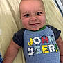baby, smile, child, face, blue_shirt, text_on_clothing, blanket, lying_down, infant, cute, happy, indoor, skin, head, eyes, short_sleeves, person, photography, closeup, playful