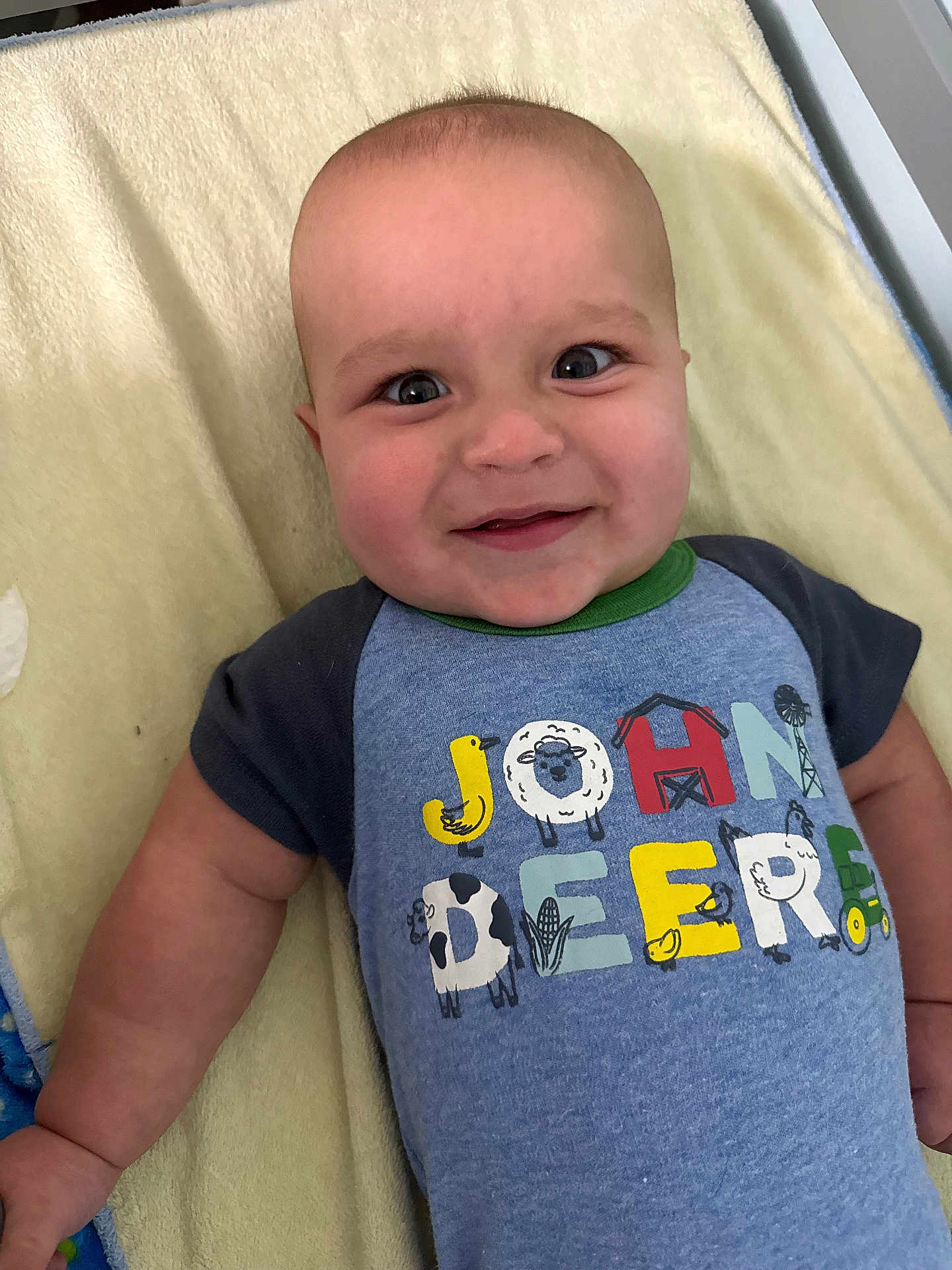Silas is registered to the contest to win money with this photo: baby, smile, child, face, blue_shirt, text_on_clothing, blanket, lying_down, infant, cute, happy, indoor, skin, head, eyes, short_sleeves, person, photography, closeup, playful