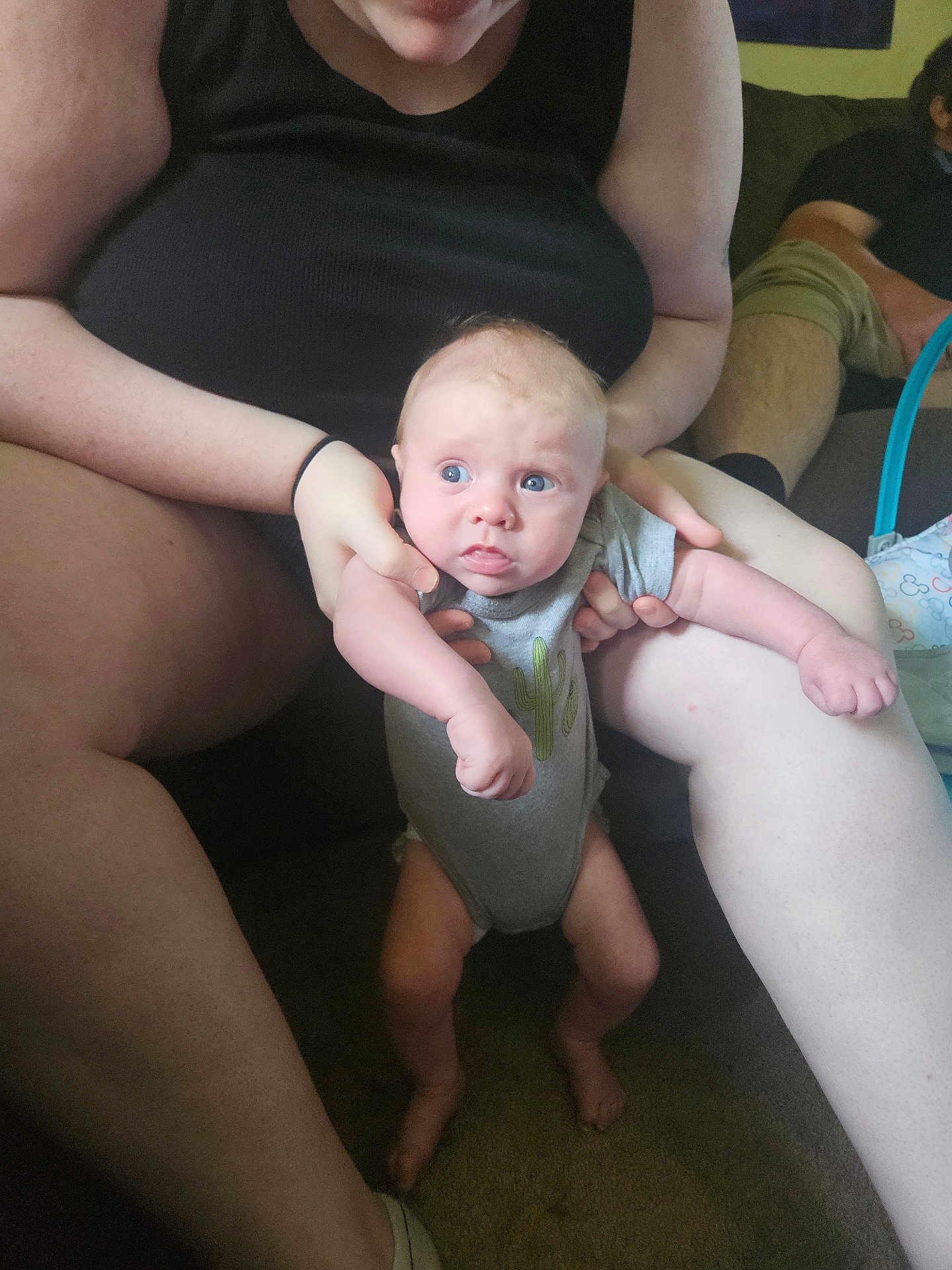Jasper is registered to the contest to win money with this photo: baby, infant, person, adult, cactus_onesie, blue_eyes, indoor, couch, carpet, sitting, holding, legs, arms, casual_clothing, relaxing, living_room, curious_expression, skin, hand, foot