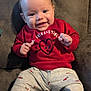 Jaxson is registered to the contest to win money with this photo: baby, child, smiling, red_sweatshirt, heart_graphic, grey_pants, arrow_pattern, sitting, couch, indoor, footwear, black_shoes, white_socks, happy, portrait, cute, infant, clothing, face, person