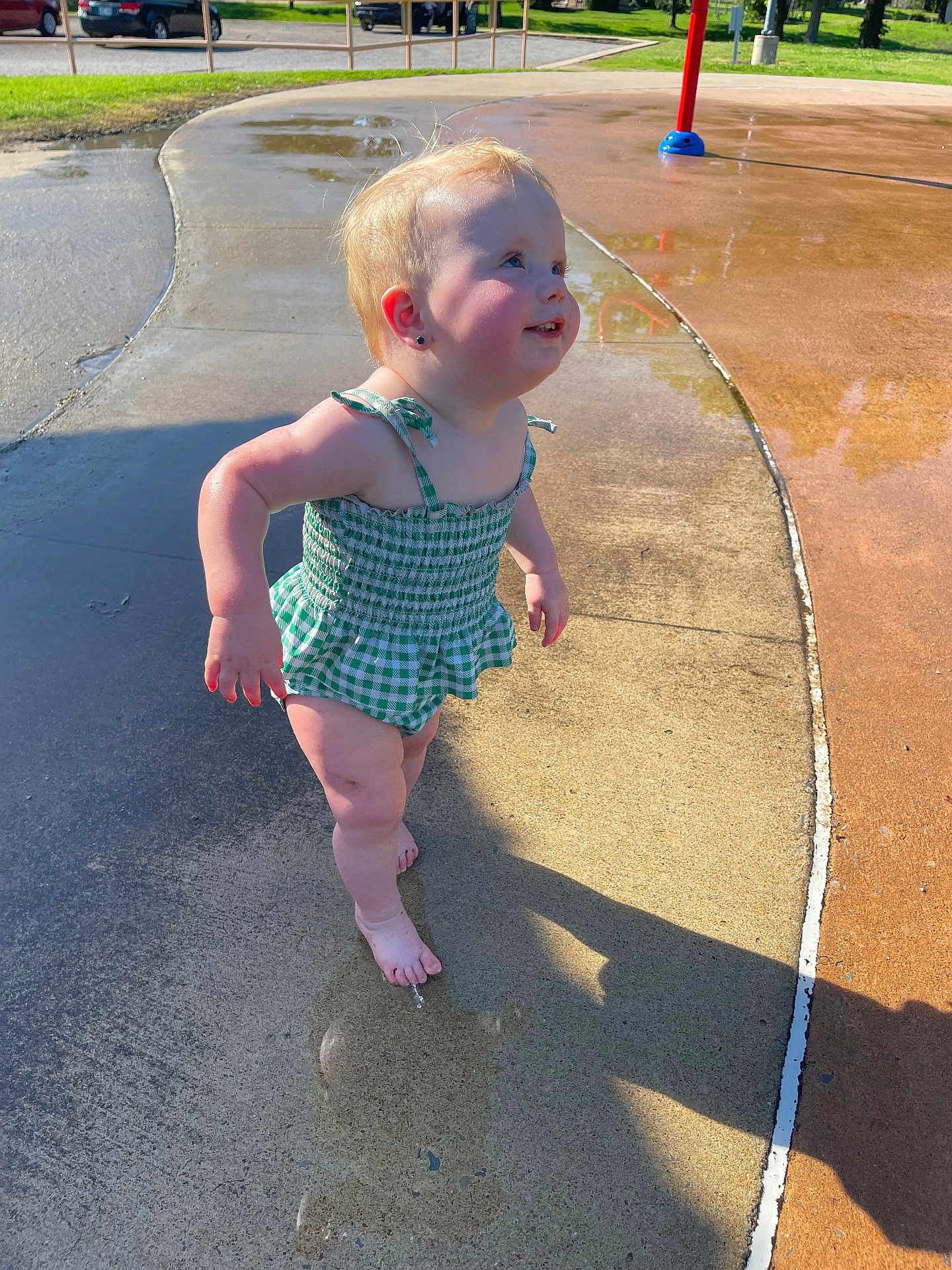 Rosey is registered to the contest to win money with this photo: toddler, child, swimsuit, green_checkered, barefoot, playground, water, wet_surface, shadow, sunlight, outdoor, summer, smile, cute, young_child, skin, earrings, concrete, park, happy