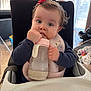 baby, bottle, bib, high_chair, food_stains, indoor, child, cute, clothing, person, hair_bow, feeding, table, seat, toy, window, natural_light, face, hand, room