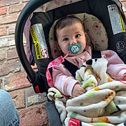 Oaklee is registered to the contest to win money with this photo: baby, pacifier, car_seat, blanket, brick_floor, jeans, slippers, child, infant, pink_clothing, seatbelt, face, indoors, person, cute, young_child, comfort, patterned_blanket, sitting, closeup