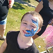 David joined the competition — help win amazing prizes! child, face_paint, blue_paint, shark_design, smile, teeth, short_hair, outdoor, grass, sunlight, black_shirt, portrait, candid, festival, event, people, hand, sandals, pink_tinsel, happy