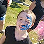 child, face_paint, blue_paint, shark_design, smile, teeth, short_hair, outdoor, grass, sunlight, black_shirt, portrait, candid, festival, event, people, hand, sandals, pink_tinsel, happy