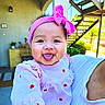 baby, child, infant, smiling, tongue_out, pink_bow, headband, pink_clothing, dress, hearts, being_held, portrait, outdoor, patio, sunlight, plants, pergola, adult, happy, playful