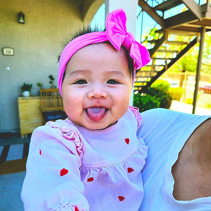 Lois Ly is registered to the contest to win money with this photo: baby, child, infant, smiling, tongue_out, pink_bow, headband, pink_clothing, dress, hearts, being_held, portrait, outdoor, patio, sunlight, plants, pergola, adult, happy, playful