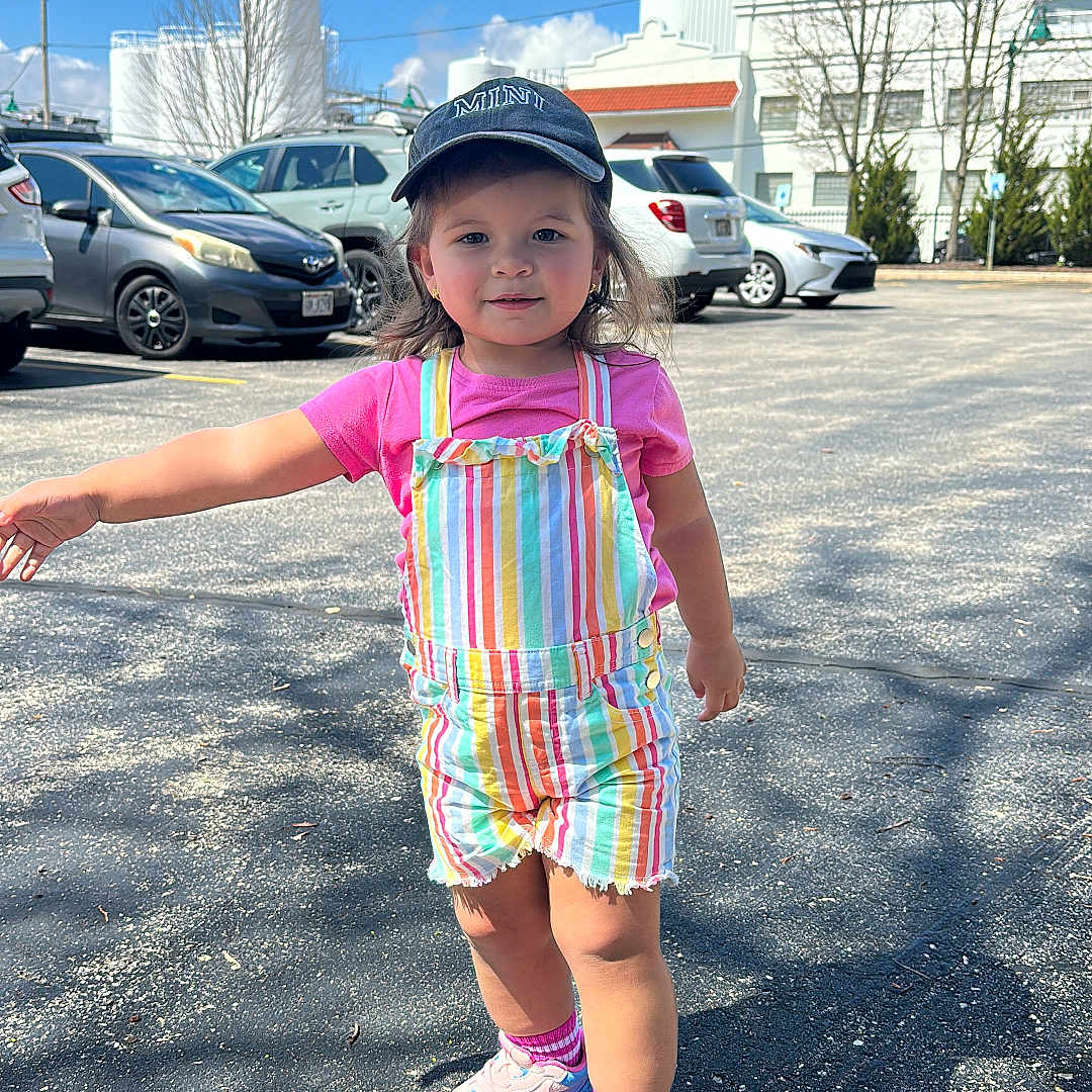 Clarette joined the competition — help win amazing prizes! toddler, child, outdoor, parking_lot, clouds, blue_sky, cap, striped_overall, pink_tshirt, sneakers, shadow, cars, asphalt, sunny, person, building, tree, daytime, smiling, casual_clothing