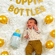 Christian joined the competition — help win amazing prizes! baby, infant, balloon, golden_balloon, baby_bottle, white_rug, banner, celebration, smiling, cute, lying_down, clothing, pants, shirt, decor, party, happy, indoors, one_person, child
