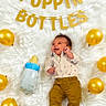 baby, infant, balloon, golden_balloon, baby_bottle, white_rug, banner, celebration, smiling, cute, lying_down, clothing, pants, shirt, decor, party, happy, indoors, one_person, child
