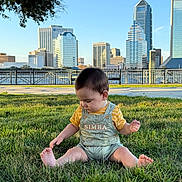 Sebástian Brown joined the competition — help win amazing prizes! baby, child, toddler, grass, outdoor, cityscape, buildings, sky, sunlight, clothing, overalls, yellow_shirt, sitting, barefoot, nature, tree, fence, daytime, playful, curious