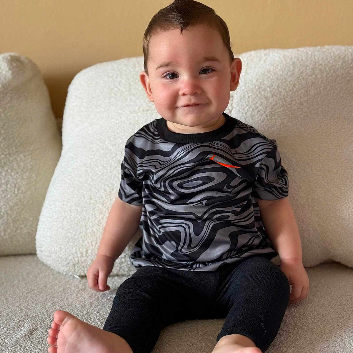 Sebástian Brown is registered to the contest to win money with this photo: barefoot, black_pants, child, clothing, couch, cozy, cushion, cute, face, hair, indoors, neutral_background, patterned_shirt, person, plush, portrait, sitting, smile, toddler, young_child