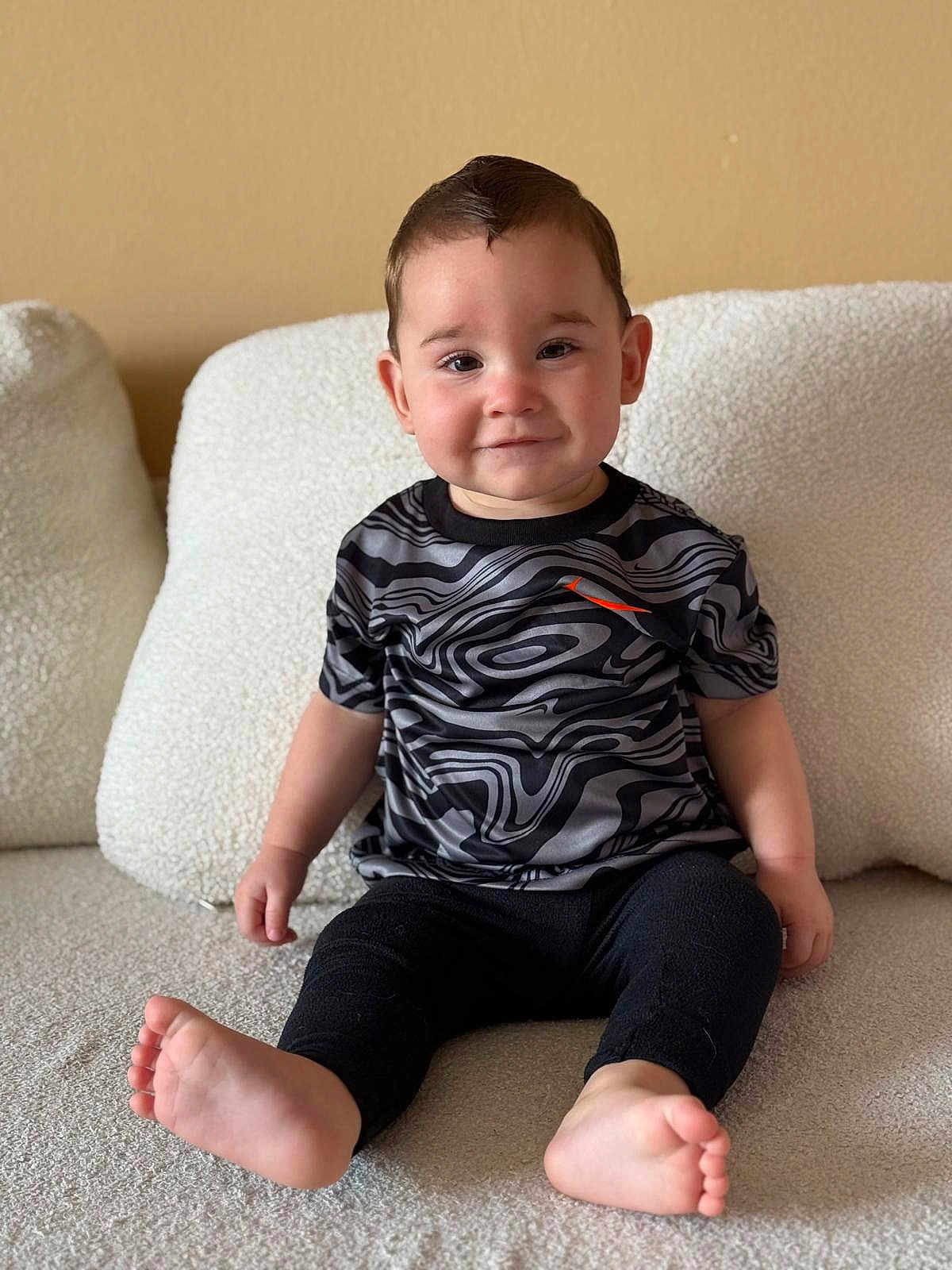 Sebástian Brown is registered to the contest to win money with this photo: toddler, child, smile, sitting, couch, clothing, black_pants, patterned_shirt, barefoot, hair, face, indoors, cozy, cushion, plush, person, cute, young_child, portrait, neutral_background