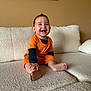baby, child, smiling, couch, orange_clothing, indoor, happy, sitting, barefoot, person, portrait, furniture, wall, artwork, home, infant, cute, toddler, cozy, hands