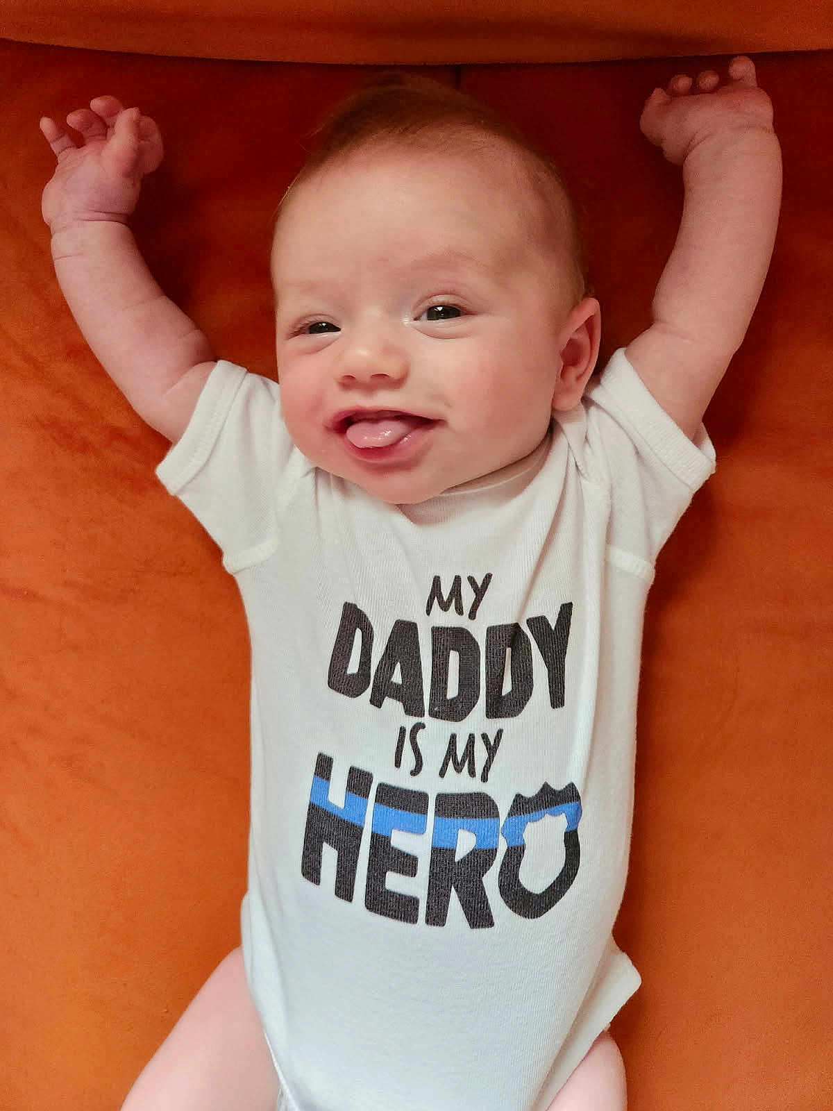 Camden joined the competition — help win amazing prizes! baby, infant, smiling, tongue_out, white_onesie, text_on_clothing, orange_background, arms_up, cute, happy, child, portrait, person, young, skin, head, face, lying_down, indoors, clothing