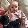 baby, infant, pink_onesie, stuffed_animal, black_plush_toy, pillow, bed, blanket, soft_texture, cuddly, cute, wide_eyes, expression, child, indoor, portrait, cozy, sleepwear, person, lying_down