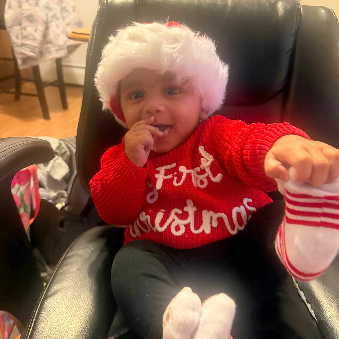 Alekzander is registered to the contest to win money with this photo: baby, black_chair, child, cozy, cute, festive, first_christmas, furniture, happy, holiday, home, indoor, person, playful, red_sweater, santa_hat, seat, smiling, striped_socks, wooden_floor