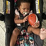 baby, car_seat, pacifier, toy, football, curly_hair, infant, child, indoors, flooring, colorful, pattern, sitting, legs, hands, face, clothing, straps, safety, playful