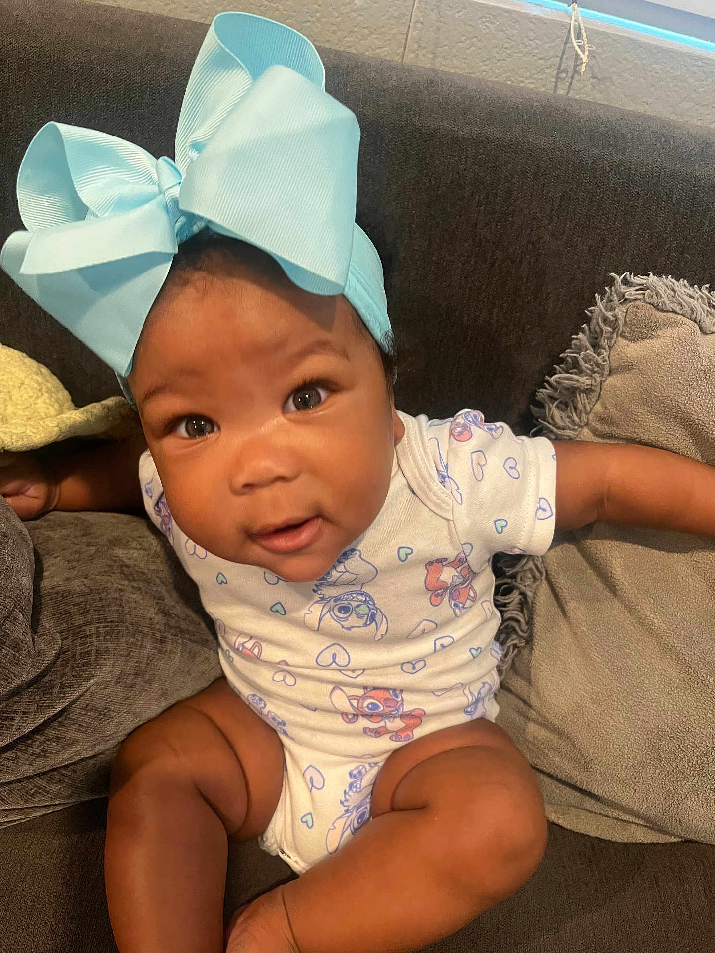 Cherish is registered to the contest to win money with this photo: baby, child, blue_bow, onesie, couch, cushion, indoor, infant, smiling, face, cute, sitting, arm, leg, headwear, clothing, portrait, happy, person, home