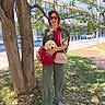 bag, building, casual_clothing, daytime, dog, grass, green_pants, holding, leaves, nature, outdoor, pet, puppy, sidewalk, smile, sunglasses, sunlight, tree, urban, woman