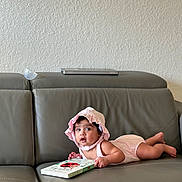Yashna joined the competition — help win amazing prizes! baby, infant, child, pink_bonnet, pink_romper, gray_couch, leather_couch, book, ladybug, reading, indoor, wall, curious, lying_down, cute, young_child, person, soft_lighting, home, portrait
