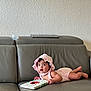 baby, infant, child, pink_bonnet, pink_romper, gray_couch, leather_couch, book, ladybug, reading, indoor, wall, curious, lying_down, cute, young_child, person, soft_lighting, home, portrait