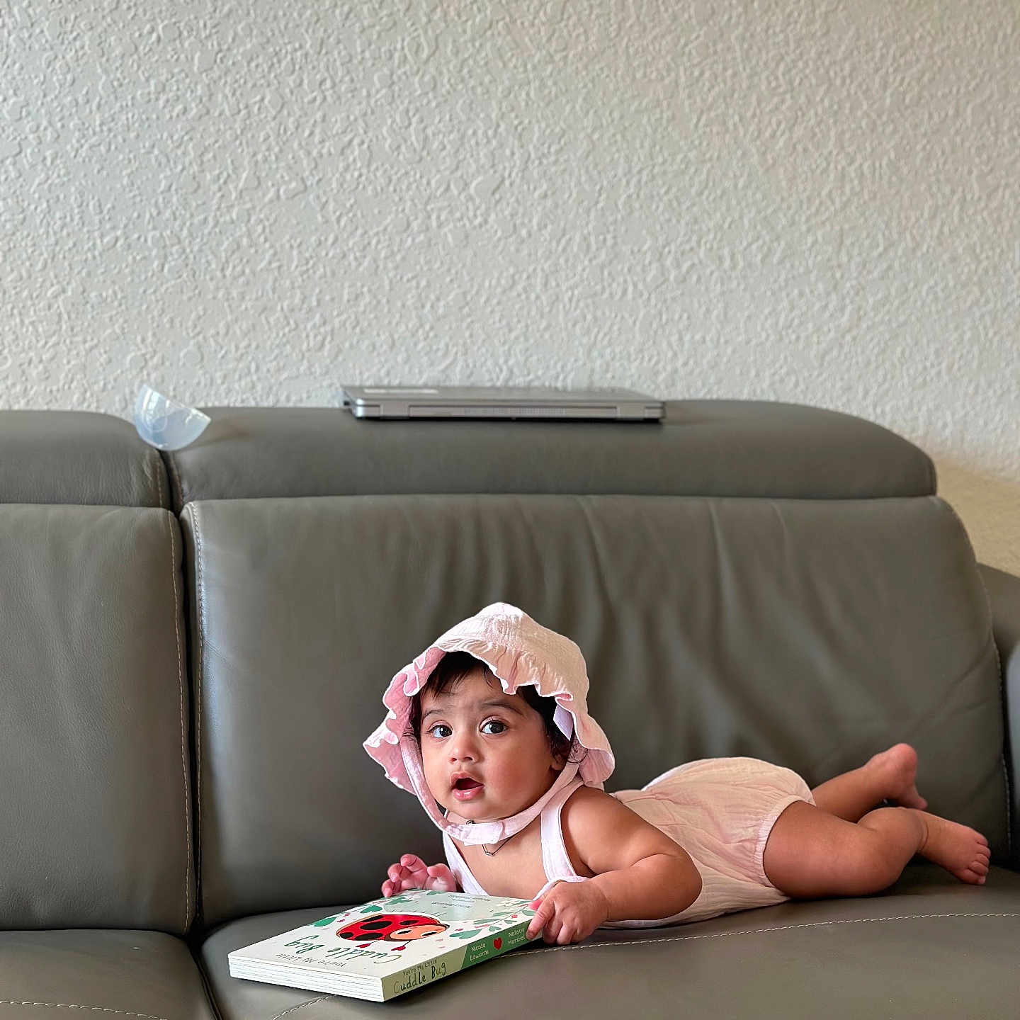 Yashna joined the competition — help win amazing prizes! baby, book, child, curious, cute, gray_couch, home, indoor, infant, ladybug, leather_couch, lying_down, person, pink_bonnet, pink_romper, portrait, reading, soft_lighting, wall, young_child