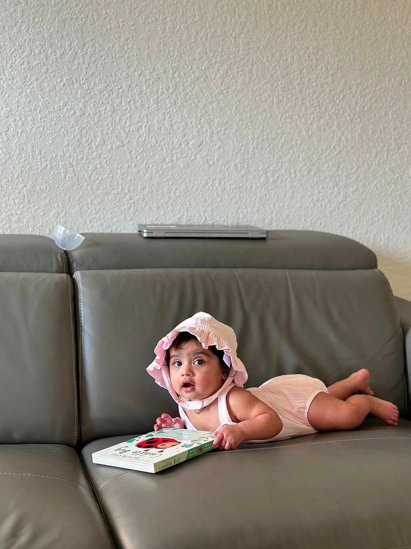 Yashna joined the competition — help win amazing prizes! baby, infant, child, pink_bonnet, pink_romper, gray_couch, leather_couch, book, ladybug, reading, indoor, wall, curious, lying_down, cute, young_child, person, soft_lighting, home, portrait