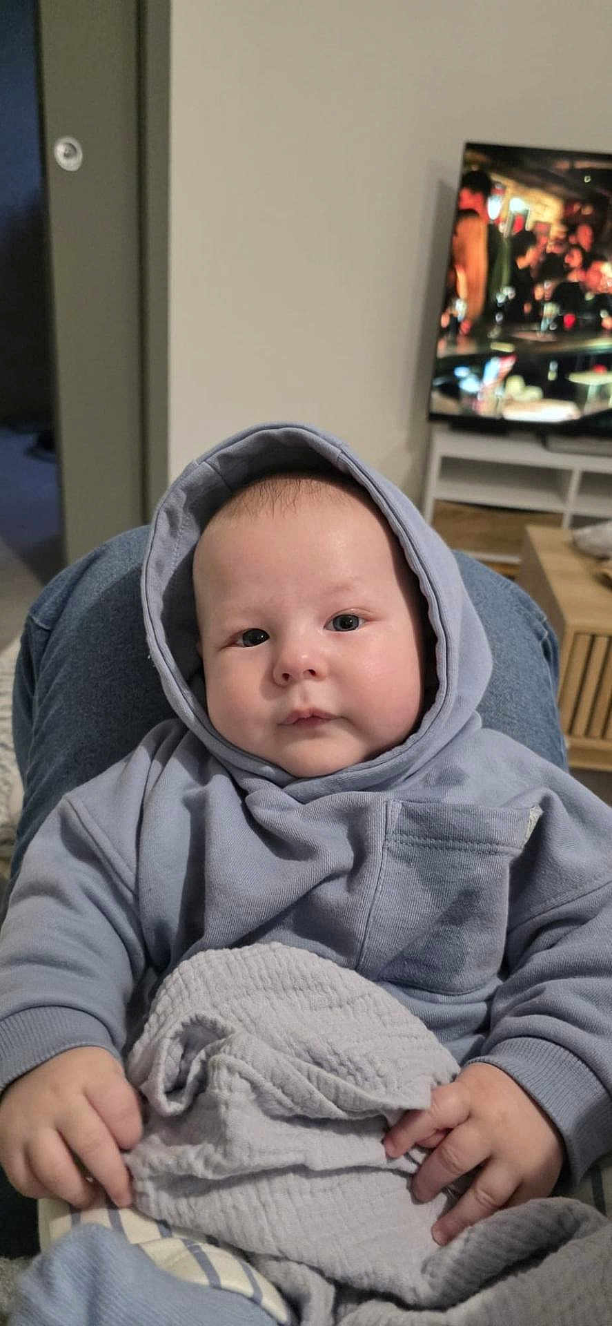 Timiarii participe au concours pour gagner de l'argent avec cette photo : baby, hoodie, blanket, indoor, child, face, hands, seated, person, television, furniture, living_room, cozy, casual_clothing, blue, soft_texture, curious, young_child, portrait, home