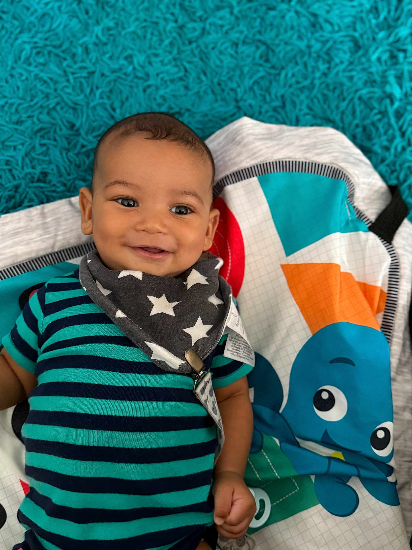 Zeke is registered to the contest to win money with this photo: baby, infant, smile, striped_shirt, bib, stars, play_mat, cartoon_octopus, blue, teal, orange, fabric, child, cute, happy, lying_down, indoors, face, person, young