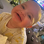 Freya is registered to the contest to win money with this photo: baby, blanket, blurred_background, cheeks, child, christmas_tree, couch, cozy, face, happy, home, indoor, infant, onesie, person, remote_control, smiling, soft_lighting, television, yellow