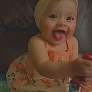 Leilah joined the competition — help win amazing prizes! baby, child, smiling, tongue_out, headband, flower, dress, sitting, couch, indoors, happy, cute, playful, person, infant, heart_emoji, colorful, young_child, portrait, fun