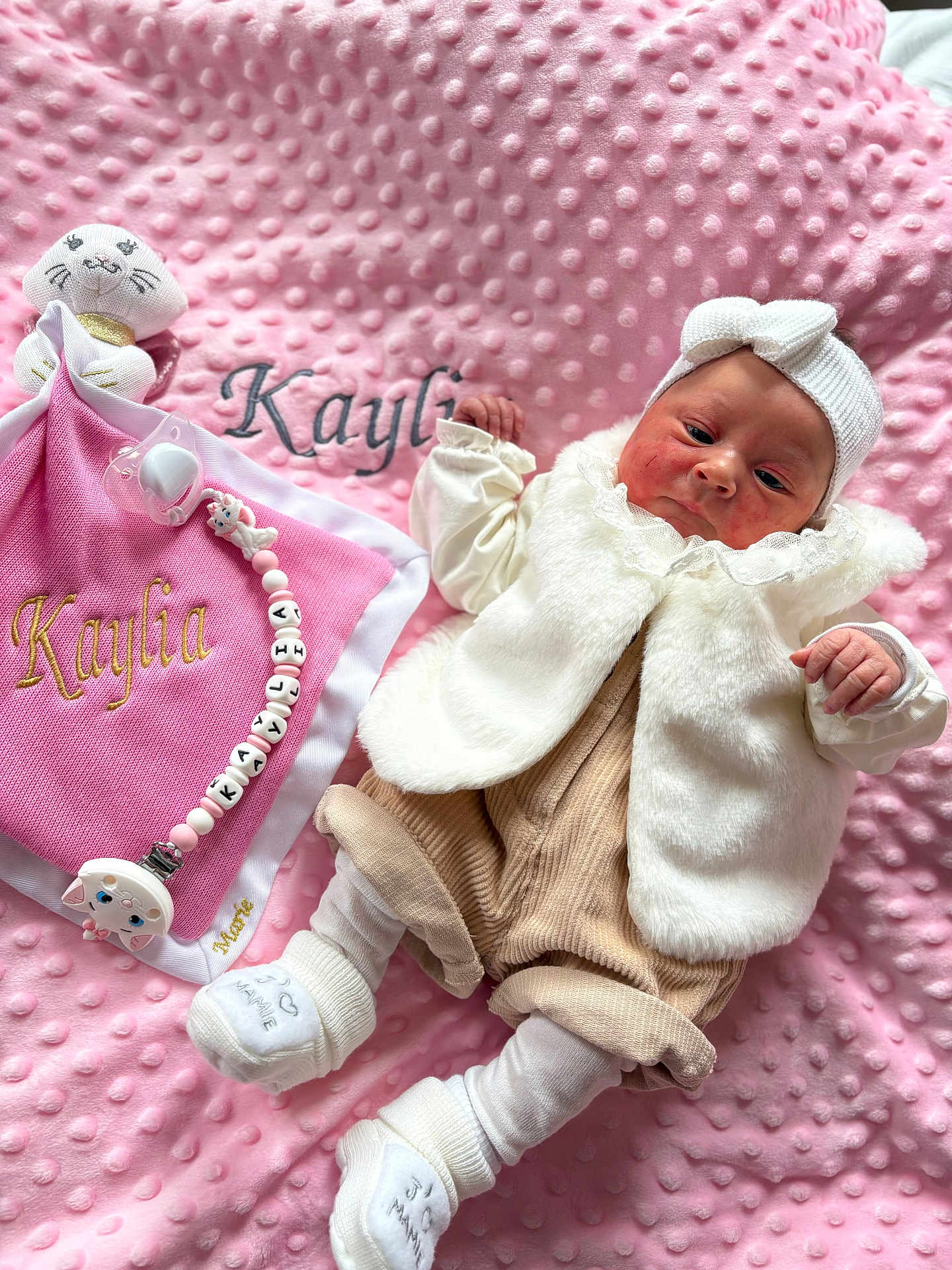 Kaylia a rejoint le concours — aidez-le/la à gagner de superbes lots ! baby, infant, pink_blanket, soft_texture, headband, white_vest, beige_pants, white_socks, pacifier, plush_toy, name_embroidery, cute, indoors, portrait, child, newborn, cozy, soft_lighting, small_hands, lying_down