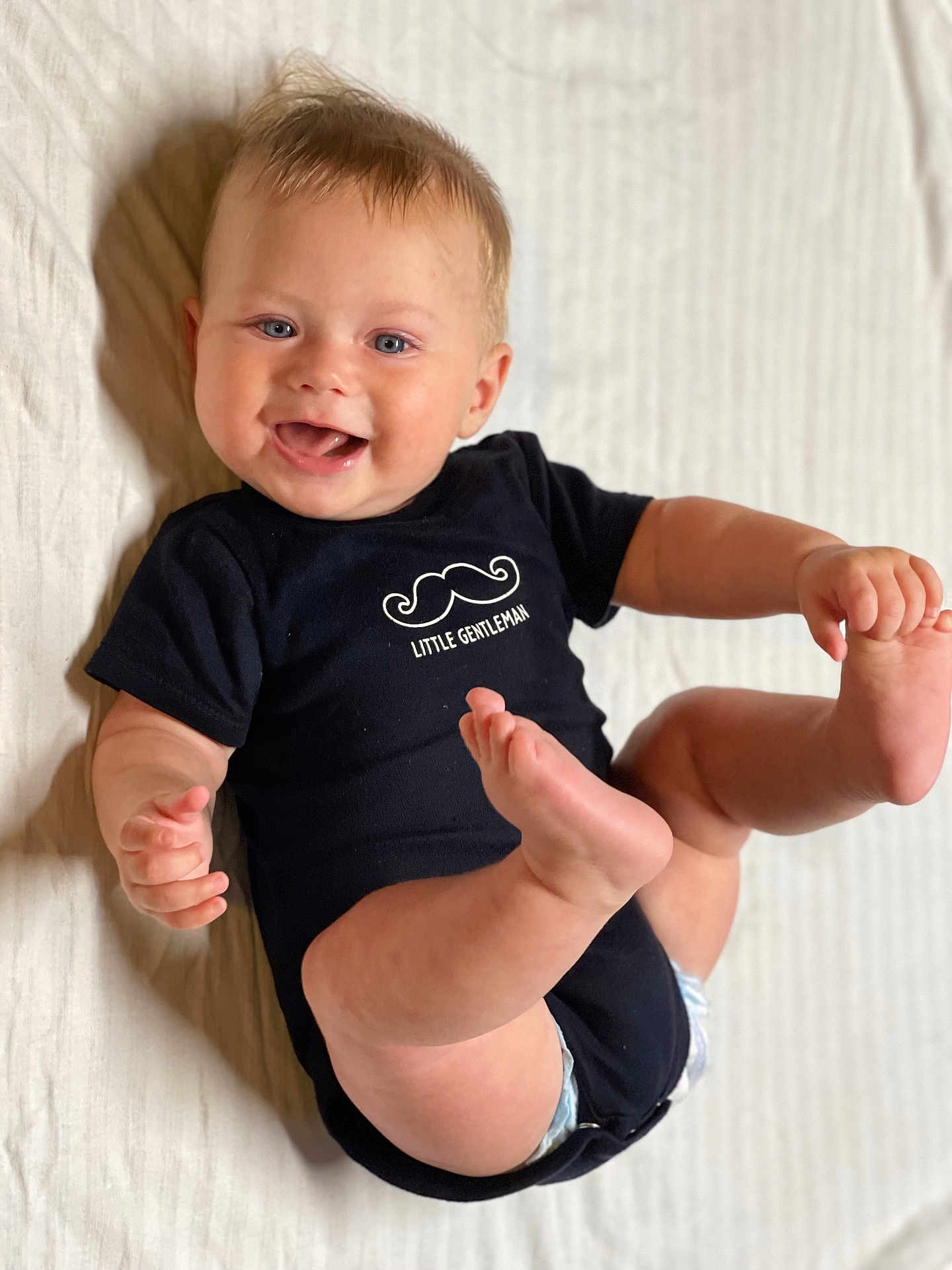 Alden joined the competition — help win amazing prizes! baby, child, infant, smiling, onesie, black_clothing, mustache_graphic, text, little_gentleman, lying_down, kicking_legs, happy, blue_eyes, short_hair, indoor, white_background, cute, diaper, skin, person