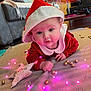 baby, infant, santa_hat, red_clothing, blanket, fairy_lights, golden_baubles, indoor, carpet, holiday, christmas, festive, cute, face, eyes, hand, soft_toys, home, decor, floor