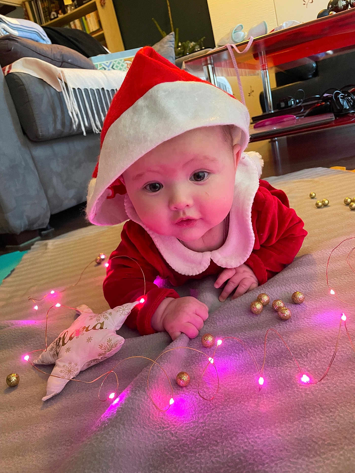 Clara a rejoint le concours — aidez-le/la à gagner de superbes lots ! baby, infant, santa_hat, red_clothing, blanket, fairy_lights, golden_baubles, indoor, carpet, holiday, christmas, festive, cute, face, eyes, hand, soft_toys, home, decor, floor
