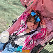 Ella'Mia is registered to the contest to win money with this photo: toddler, stroller, sunglasses, pink, plaid_jacket, jeans, baby_bottle, pacifier, child, outdoor, sunlight, reflection, casual_clothing, seatbelt, curly_hair, relaxed, playful, cute, baby_accessories, infant
