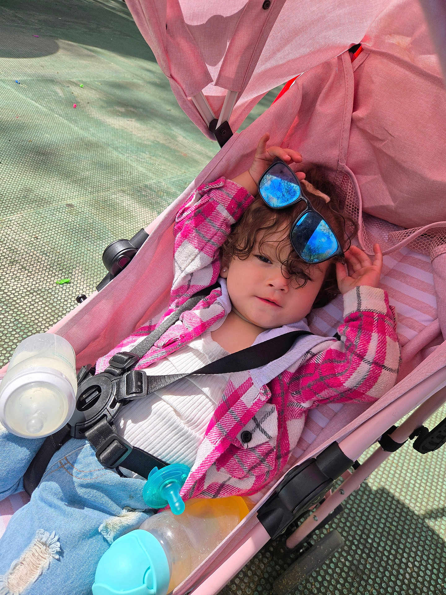 Ella'Mia is registered to the contest to win money with this photo: toddler, stroller, sunglasses, pink, plaid_jacket, jeans, baby_bottle, pacifier, child, outdoor, sunlight, reflection, casual_clothing, seatbelt, curly_hair, relaxed, playful, cute, baby_accessories, infant
