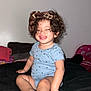 toddler, child, smile, curly_hair, headband, blue_onesie, sitting, barefoot, indoor, black_bedspread, happy, cute, baby, person, skin, face, hand, foot, toes, clothing