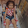 toddler, child, stroller, indoor, curly_hair, polka_dot, shirt, sandals, doll, blanket, floor, metallic_appliance, seatbelt, curious, young_child, person, baby, footwear, toy, casual