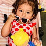 toddler, child, curly_hair, sunglasses, polka_dot, red, pink, doll, toy, stroller, seatbelt, clothing, portrait, indoor, cute, baby, playful, holding, person, young_child