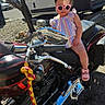 toddler, child, motorcycle, sunglasses, outdoor, sunny, tree, van, pink_shoes, purple_bows, striped_dress, vehicle, handlebars, chrome, road, rock, shadow, person, summer, casual
